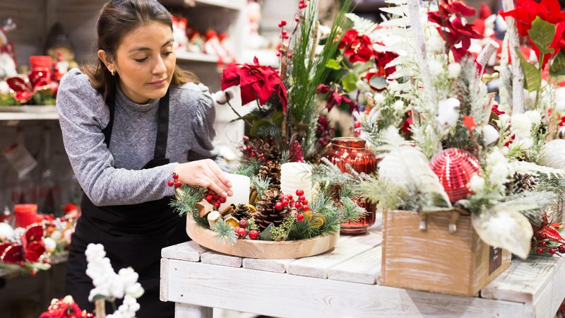 A woman arranges Christmas decorations on a white wooden table amid festive items like pine cones, berries, and red and white ornaments.