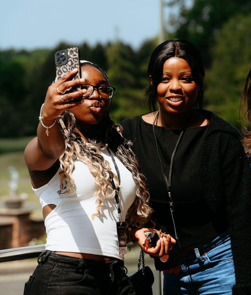 Two young women are taking a selfie outdoors, with trees and a fountain in the background, both smiling and dressed casually in black and white.
