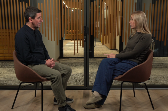 A man and woman sit facing each other in a room with wooden walls and glass doors behind them, engaged in a conversation.