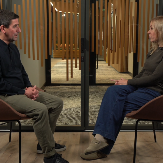 A man and woman sit facing each other in a room with wooden walls and glass doors behind them, engaged in a conversation.