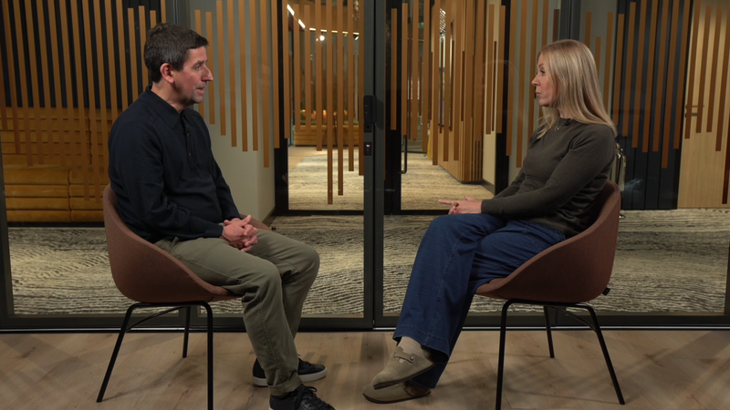 A man and woman sit facing each other in a room with wooden walls and glass doors behind them, engaged in a conversation.