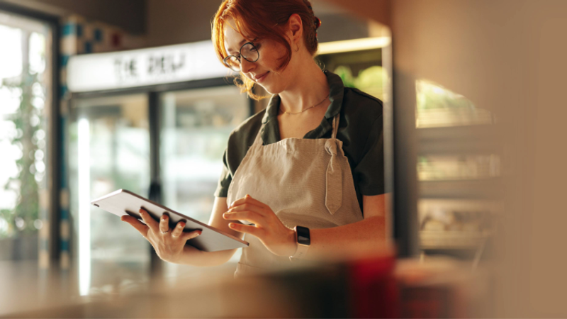 A woman with red hair and glasses, wearing a black shirt and apron, is looking at a tablet in a well-lit café or shop interior.