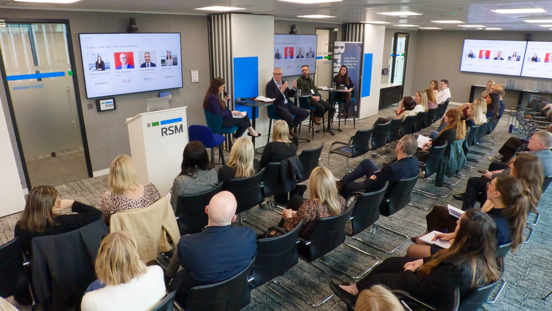 A panel discussion taking place in a modern conference room with an audience of around 25 people, alongside screens displaying the panel topics and speakers.