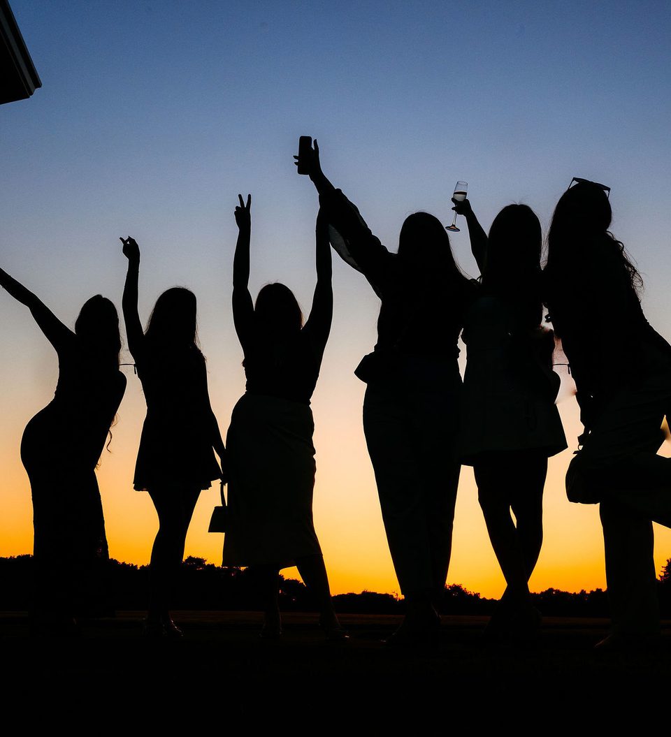 Six women are standing together outdoors during sunset, some raising their arms and holding a glass, with a railing and trees visible in the background.