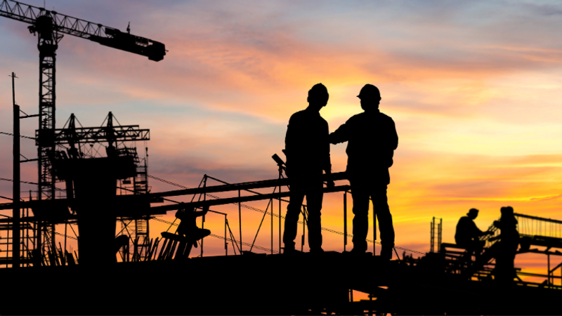 Silhouettes of construction workers on a scaffolded platform at sunset, with cranes and machinery against a warm orange sky.