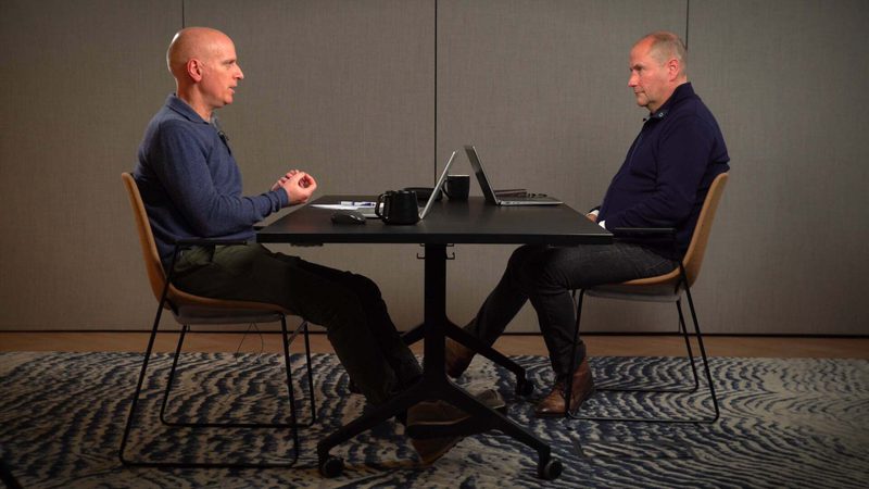 Two men sit facing each other at a small black table, with laptops and coffee mugs, in a neutral-toned room.