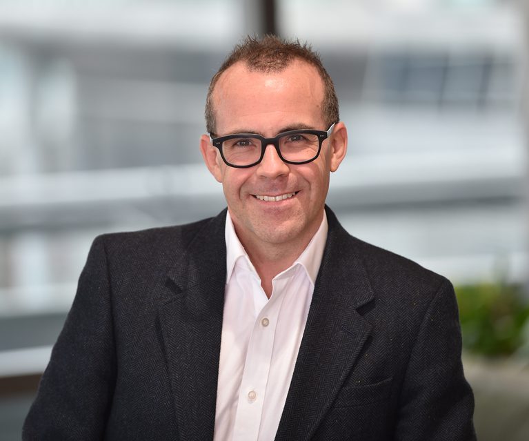A man with short brown hair, wearing black glasses, a white shirt, and a dark blazer, smiling in an indoor modern office environment.