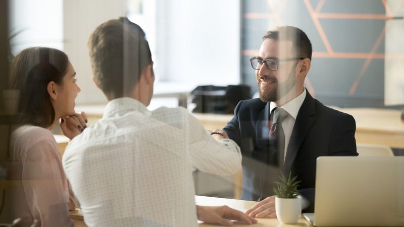 A professional man in a suit and glasses smiling during a meeting with two colleagues, a woman and a man, behind a glass partition in an office.