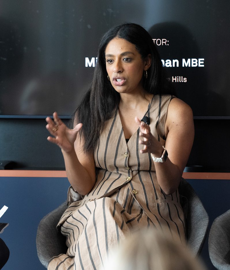A woman with long dark hair, wearing a sleeveless beige and black striped dress, speaking and gesturing with her hands during a panel discussion.