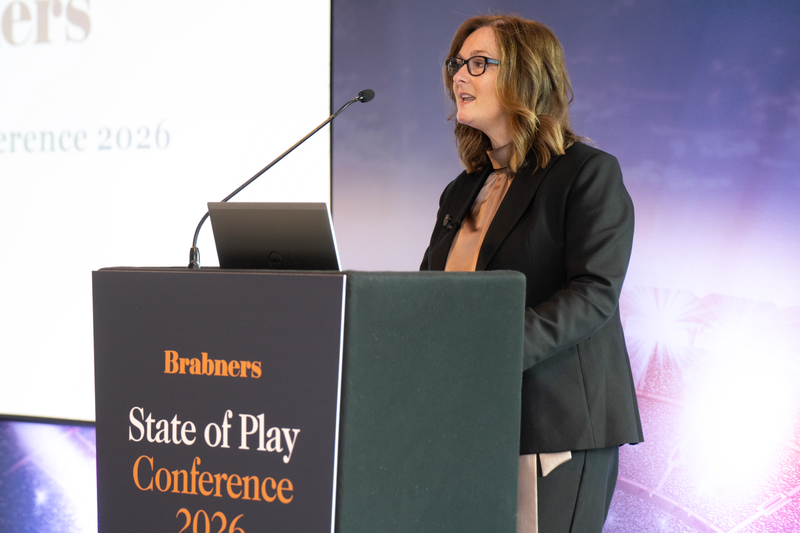 A speaker wearing a dark blazer and glasses addresses the audience from a podium with a laptop and microphone at the Brabners State of Play Conference 2026.