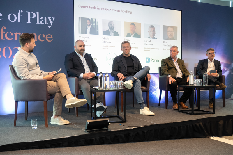 Five panelists sit on stage at a conference, with water bottles on side tables; a large screen behind displays portraits and logos for sport tech in event hosting.