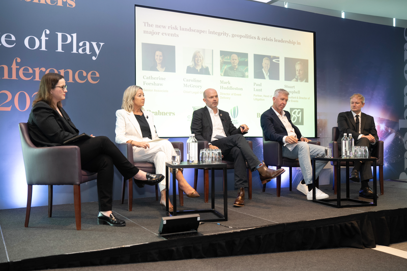 Five panelists seated on stage at a conference, with a large screen showing headshots and agenda; water bottles on tables against a blue backdrop.