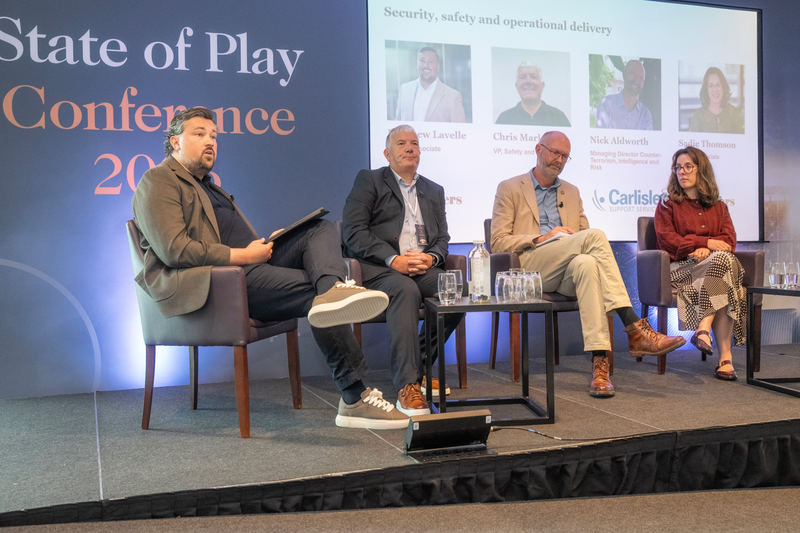 Four panelists sit on a stage at a conference, with a large blue backdrop reading State of Play Conference. A screen shows speakers and logos; water glasses on a table.