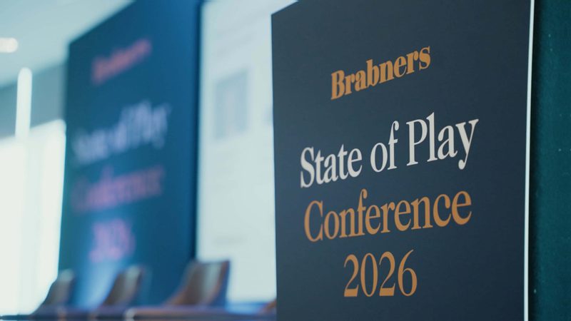 Blue conference sign reading Brabners State of Play Conference 2026 in white and orange, with a blurred room and chairs in the background