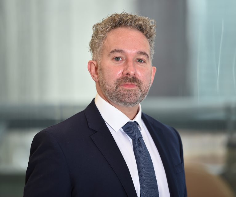 A middle-aged man with curly, light-colored hair and a beard, wearing a dark suit, white shirt, and a dark tie, standing in a modern office environment.