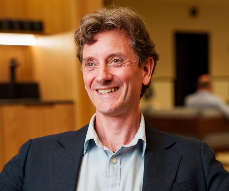 Smiling person with wavy grey hair, wearing a light blue shirt and dark blazer, in a warmly lit indoor space with wooden panels and a blurred background.