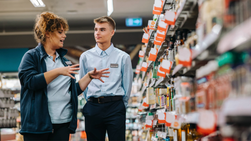 A customer and a shop assistant are talking in a store aisle filled with shelves of products, with the customer gesturing as they speak.