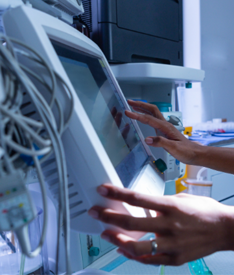 A healthcare professional in blue scrubs and a stethoscope operates a touchscreen medical monitor in a hospital, with tubes, cables and other equipment nearby, appearing to adjust settings.