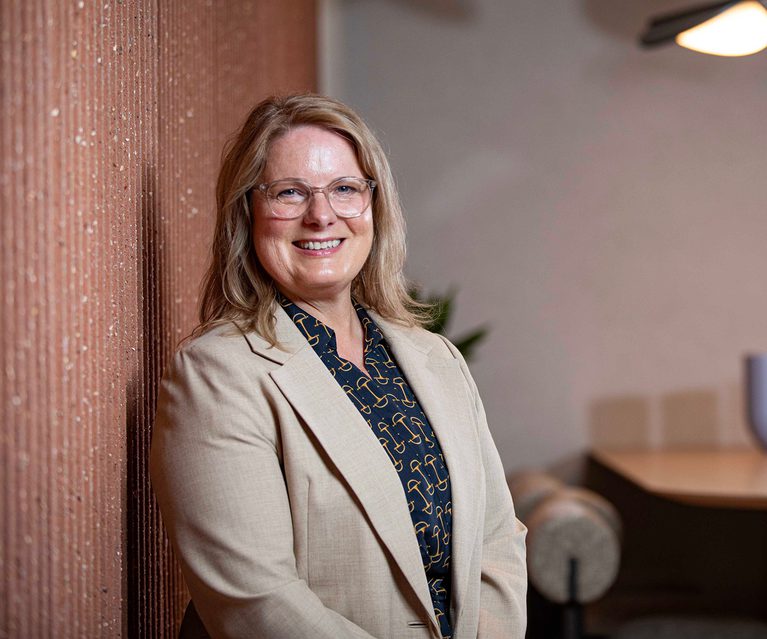 Smiling person with shoulder-length blonde hair and glasses, wearing a beige blazer over a navy patterned shirt, leaning against a textured brick wall indoors.