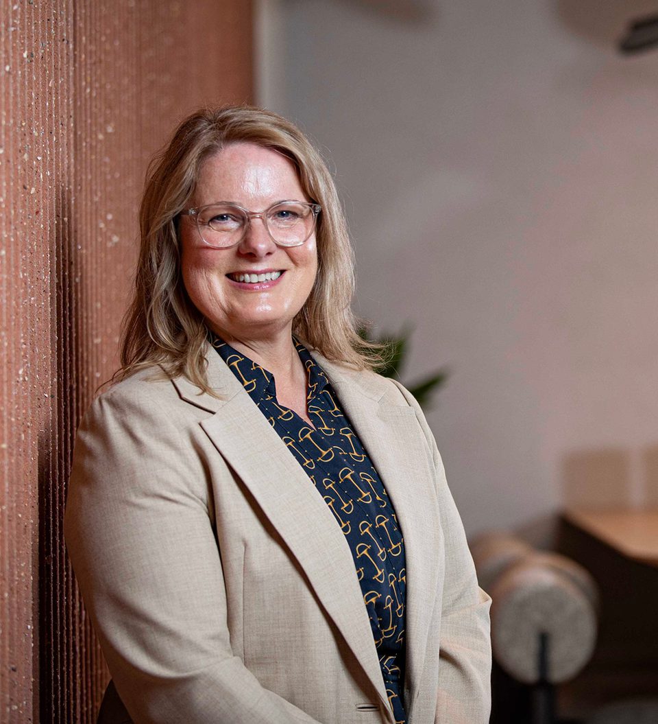 Smiling person with shoulder-length blonde hair and glasses, wearing a beige blazer over a navy patterned shirt, leaning against a textured brick wall indoors.