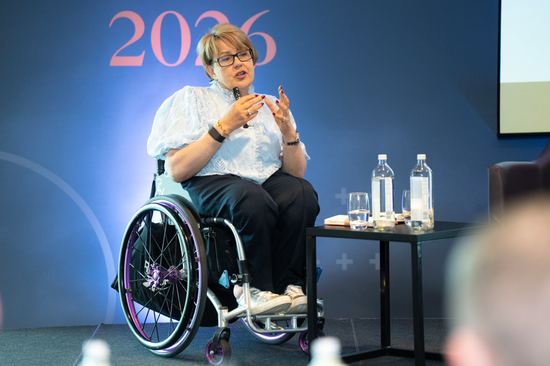 A person in a wheelchair speaks on a blue stage with a pink 2026 backdrop, wearing a light blue patterned blouse and glasses, gesturing with hands beside a water bottle table.