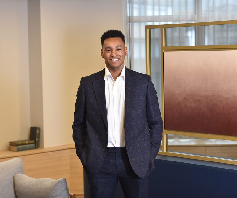 A smiling man in a dark checkered suit with a white shirt stands indoors with his hands in his pockets, in front of a display panel and bookshelf.