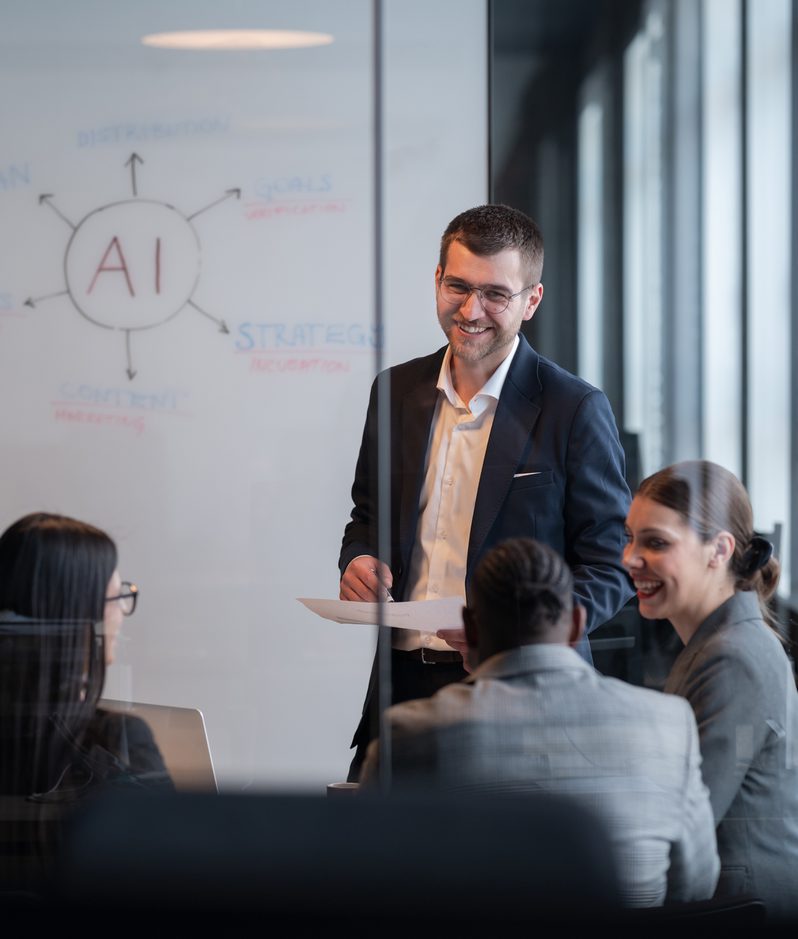 A man in a suit stands smiling with a tablet, presenting to three women seated at a table in a modern office with a whiteboard marked with "AI" and related terms.