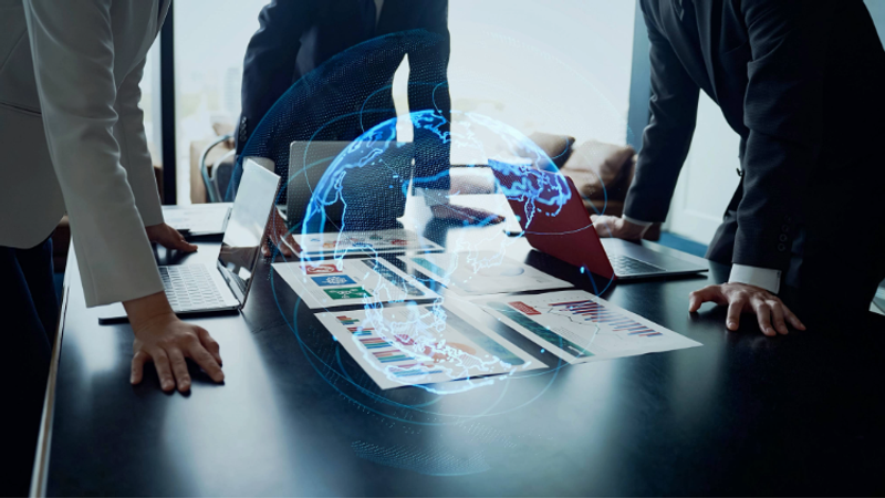 People gathered around a table with laptops and documents, overlaid with a digital globe and data streams, suggesting a technology or business meeting.