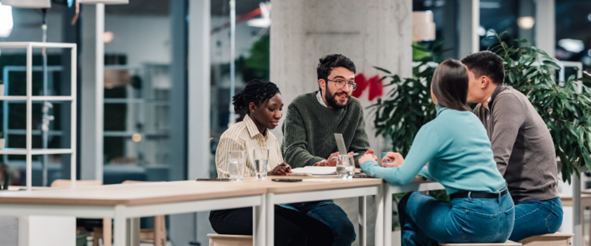 Four people sit around a high table in a modern office, collaborating over papers and laptops, with plants and warm lighting in the background.