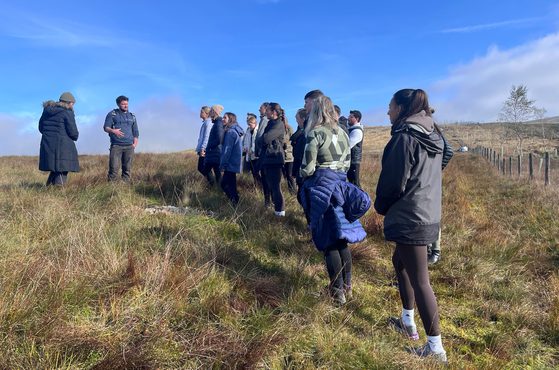 A group of people standing on a grassy hillside, listening to a person who appears to be giving a talk or guided tour, with a blue sky and some clouds overhead.