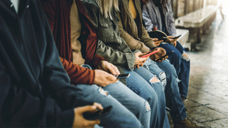 A row of people seated on a bench along a canal walkway, each focused on their smartphones, wearing casual jackets and ripped jeans as daylight glints off wet pavement.