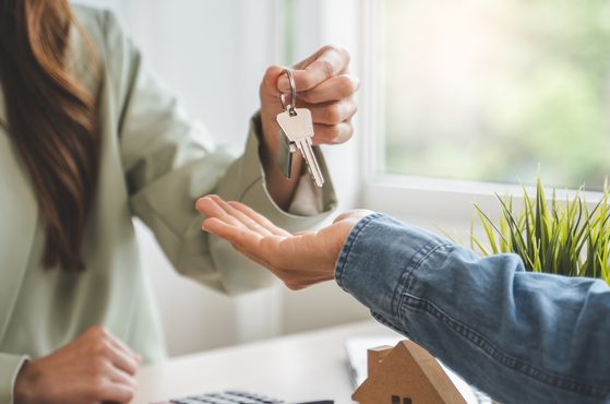 A person with long hair hands over a set of keys to another individual inside a bright room near a window with greenery outside.
