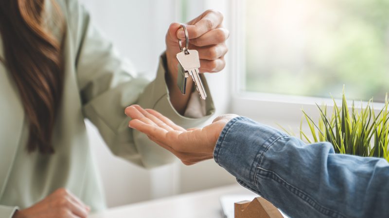 A person with long hair hands over a set of keys to another individual inside a bright room near a window with greenery outside.