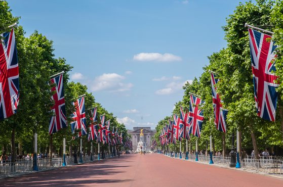 Sunlit avenue lined with Union Jack flags and lush trees, red-paved road, crowds behind barriers, and a distant historic building at the end under a blue sky.