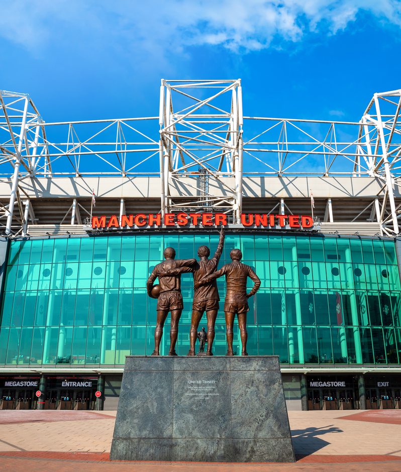 Bronze statue of three football players with arms around each other, in front of Manchester United's glass-fronted stadium beneath a bright blue sky.
