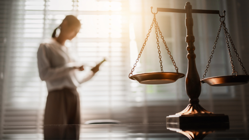 A blurred figure in a bright office holds a tablet, while a large set of scales of justice sits prominently on a polished desk in the foreground.