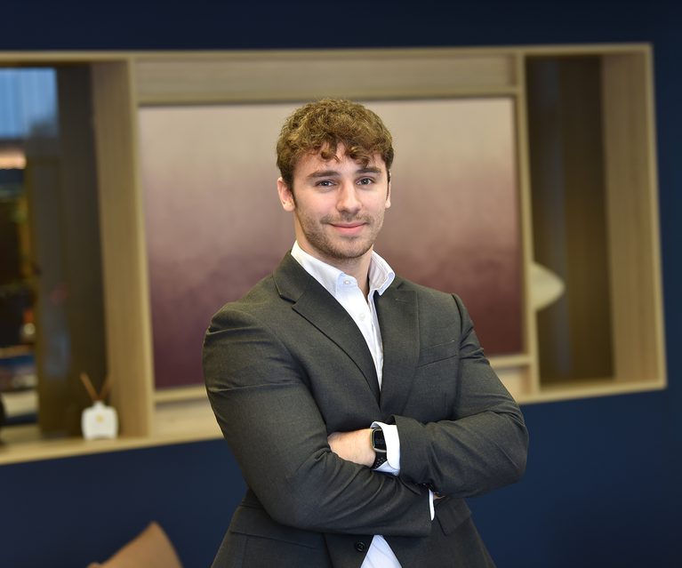 A young man with curly brown hair, wearing a dark suit and white shirt, smiling with arms crossed, standing indoors in front of a wooden shelf with a blue background.