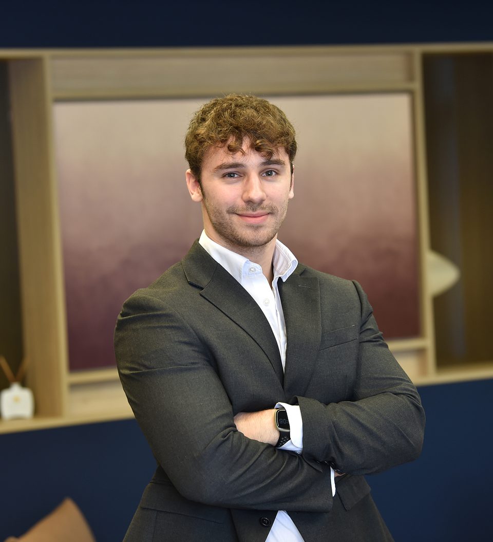 A young man with curly brown hair, wearing a dark suit and white shirt, smiling with arms crossed, standing indoors in front of a wooden shelf with a blue background.