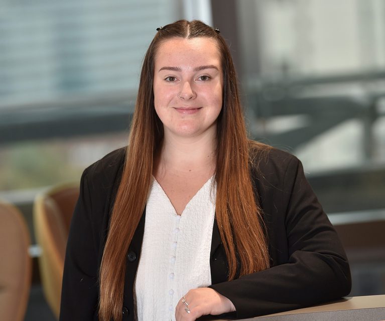 A young woman with long, straight auburn hair, wearing a black blazer over a white blouse, smiling and seated at a table in a modern office setting.