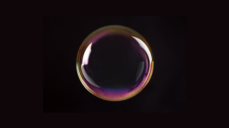A single iridescent soap bubble on a dark background, its rainbow film displaying purple and gold hues with a bright reflective highlight.