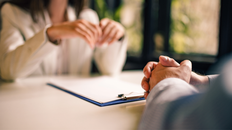 Two people sit at a table; foreground shows clasped hands, while the background person gestures with their hands; a blue clipboard rests on papers.