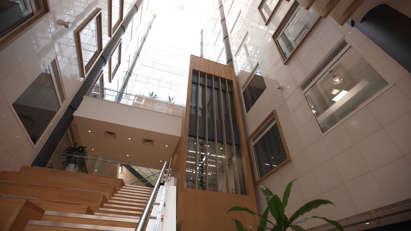 A modern indoor atrium with a glass ceiling, featuring wooden staircase, framed windows on walls, and a central glass-enclosed elevator shaft.