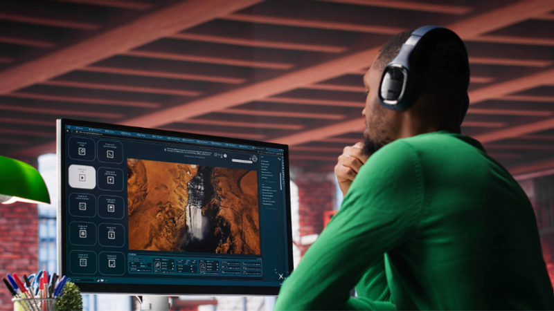 A man wearing a green shirt and headphones is working at a desk, analysing images of a waterfall on a large computer monitor in a modern, industrial-style office.