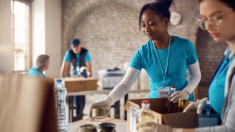 Two women in blue shirts and gloves are packing food items into boxes on a table in a bright room with brick walls and large windows.