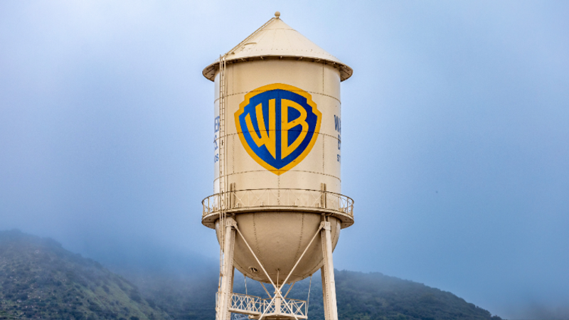 A beige cylindrical water tower with the blue and yellow Warner Bros. logo, set against a cloudy sky and misty mountains in the background.