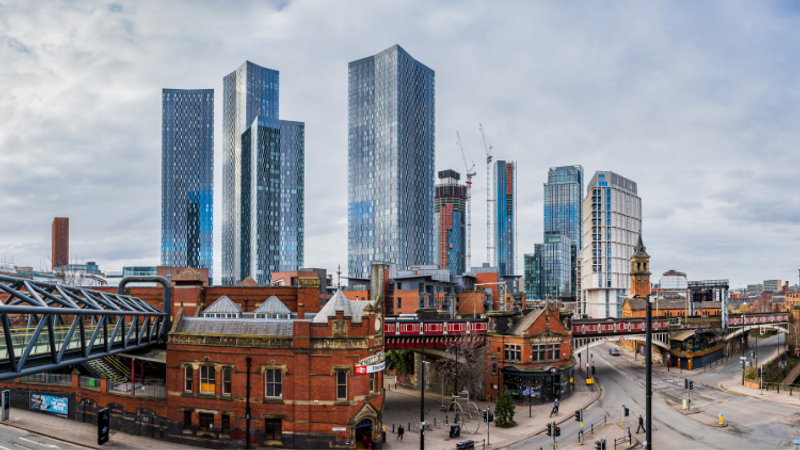 Panoramic cityscape featuring red brick historic buildings and a railway viaduct in the foreground, with a cluster of tall glass skyscrapers rising behind under a cloudy sky