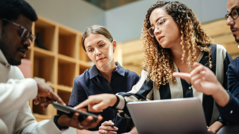 Group of four diverse people focused on a smartphone, gathered around a laptop in a modern, brightly lit workspace with wooden shelves in the background.