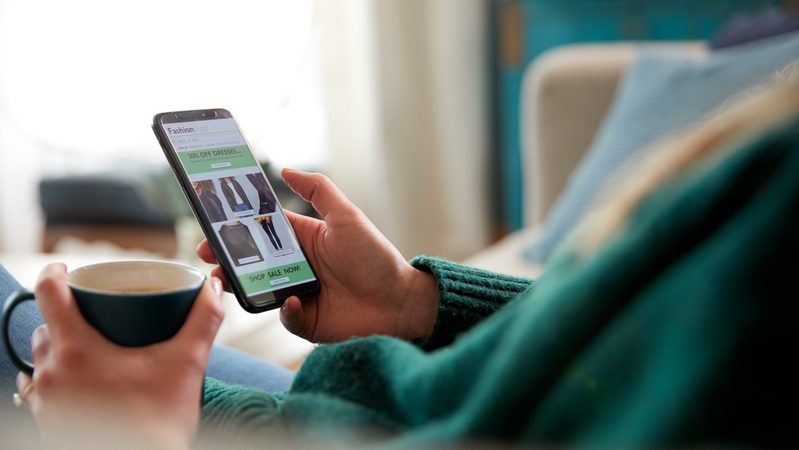 Person holding a smartphone displaying an online clothing sale, with a cup of coffee in their other hand in a cosy living room setting.