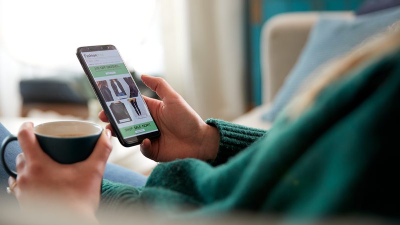 Person holding a smartphone displaying an online clothing sale, with a cup of coffee in their other hand in a cosy living room setting.
