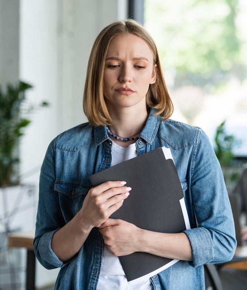 A young woman with shoulder-length blonde hair looks concerned, holding a black folder close to her chest, while a man in a suit sits at a desk in the background.